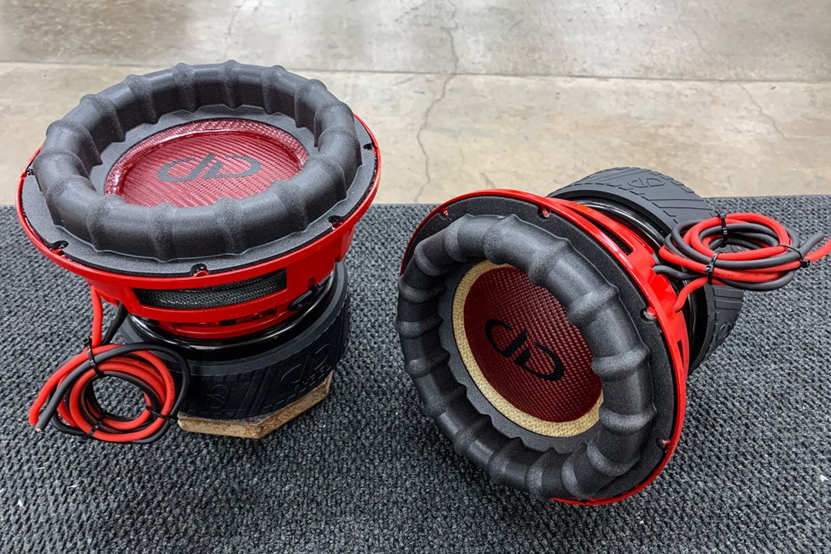 Two USA Made Subwoofers, one with red carbon fiber cone and dust cap, black DDA logo decal and the other with yellow cone, both with red powder coat frames